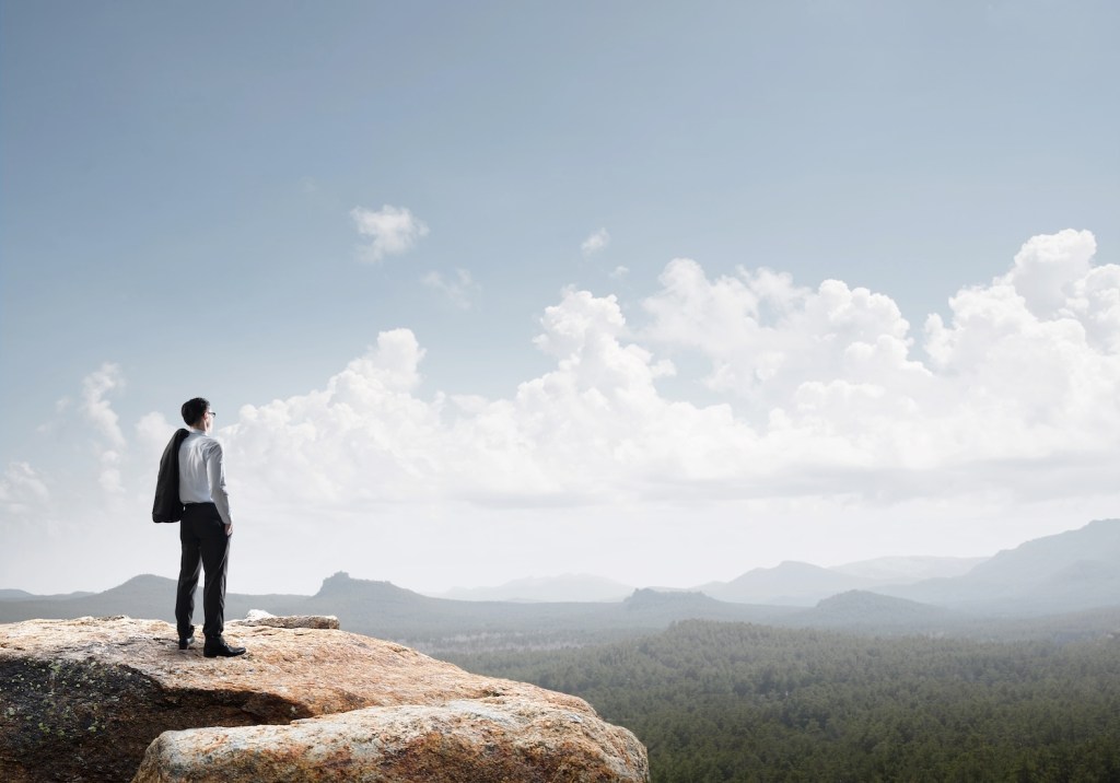 A distant adult businessperson stands on a rocky hilltop, gazing out over a landscape of green forests and distant mountains. Business success concept. Fluffy white clouds dot a pale blue sky. Concept of business ancient intelligence and power of strategic business mentoring.