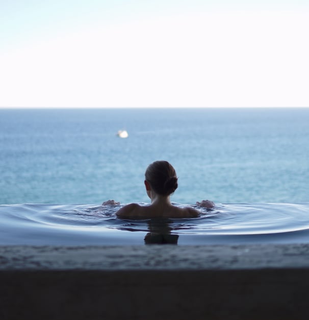 A woman in an ayurvedic resort infinity pool looking away from the viewer into the sea horizon. Concept of modern ayurvedic Gyana practice, ancient sciences integration into wellness and mindfullness.
