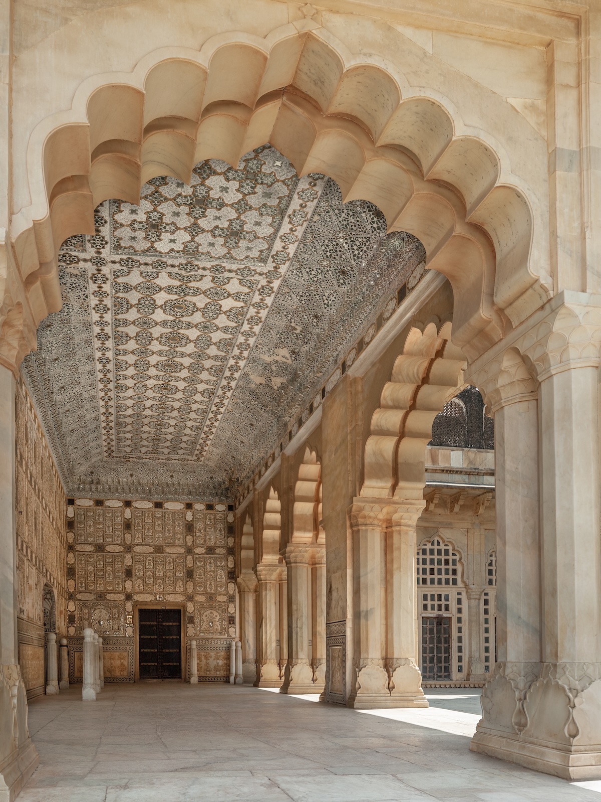 Ornate palace hallway. The hall features a series of arched entrances supported by pillars, leading deeper into the building. The ceilings and walls are elaborately decorated with intricate patterns and carvings in cream and beige tones. Soft, natural light bathes the space highlighting the light of ancient knowledge applied in modern life.