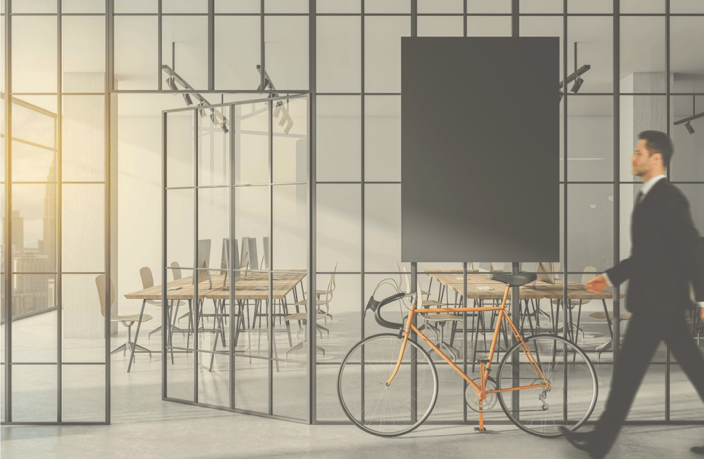 In a bright, modern office space, a man with short, light brown hair is walking in blurred motion wearing a black suit and tie. A gold-colored bicycle is in the foreground. Behind the man and bicycle is a large gray canvas. The office has walls of gray-framed glass