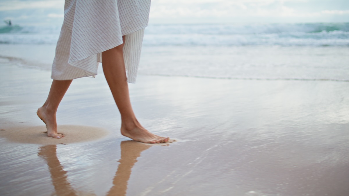 A closeup on a feet of a woman in a light-colored, striped dress who strolls barefoot on a wet, sandy beach. Concept of a relaxed knowledge Gyana community that promotes ancient vedic knowledge applied to modern luxury lifestyle.