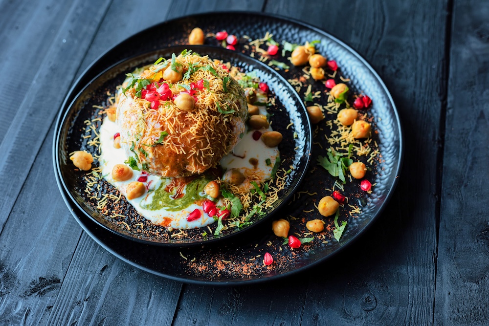 Indian chaat dish beautifully arranged on two black textured plates over a dark wooden surface. The chaat is topped with chickpeas, pomegranate seeds, herbs, and fried vermicelli. The dish is drizzled with swirls of creamy white yogurt and vibrant green chutney. Concept of masala with all six tastes as per Ayurveda