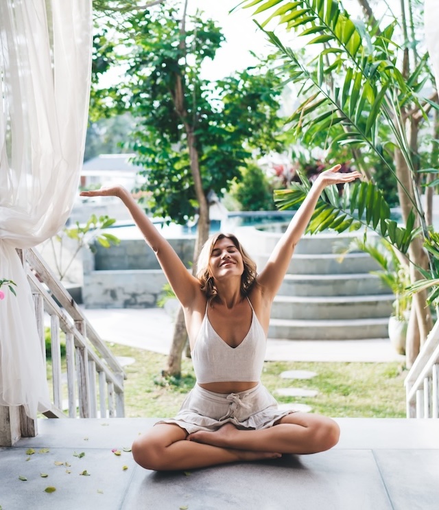 Young smiling woman with hands in air sitting on steps of her house outdoor enjoying life. Concept of vastu for home and vastu remedies assisting in personal and business life.