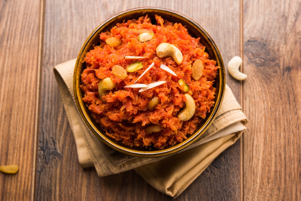 A bowl of bright orange carrot halwa topped with cashews, raisins, and almond slivers, placed on a folded beige cloth on a wooden surface. Concept of Ayurvedic use of carrots.