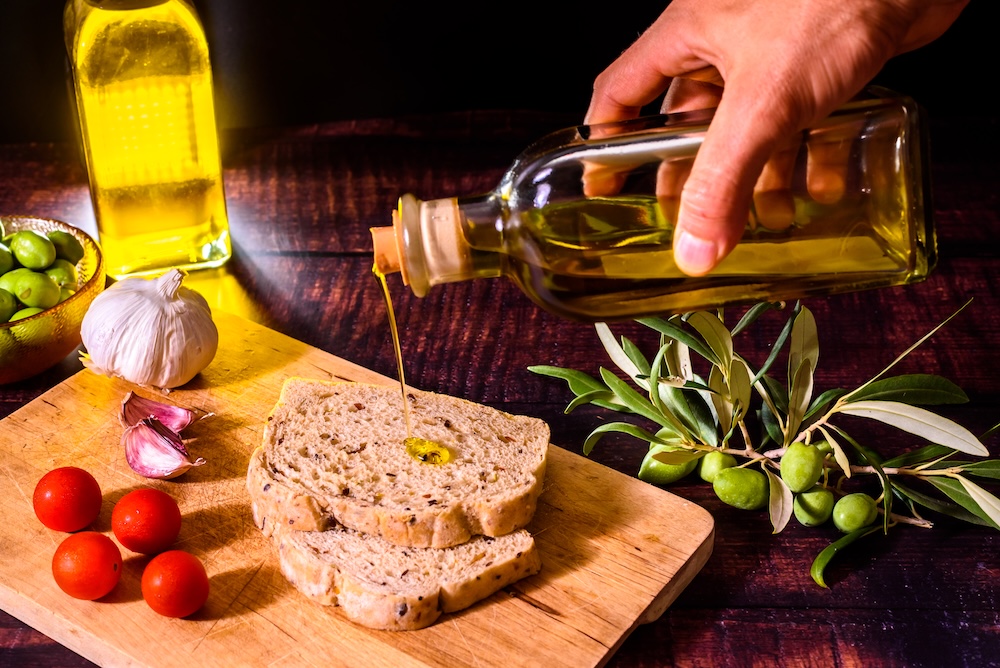 Person's hand drops a stream of extra virgin olive oil on a slice of bread. Ayurvedic uses and properties of olive oil concept, how to find out if olive oil is fake.